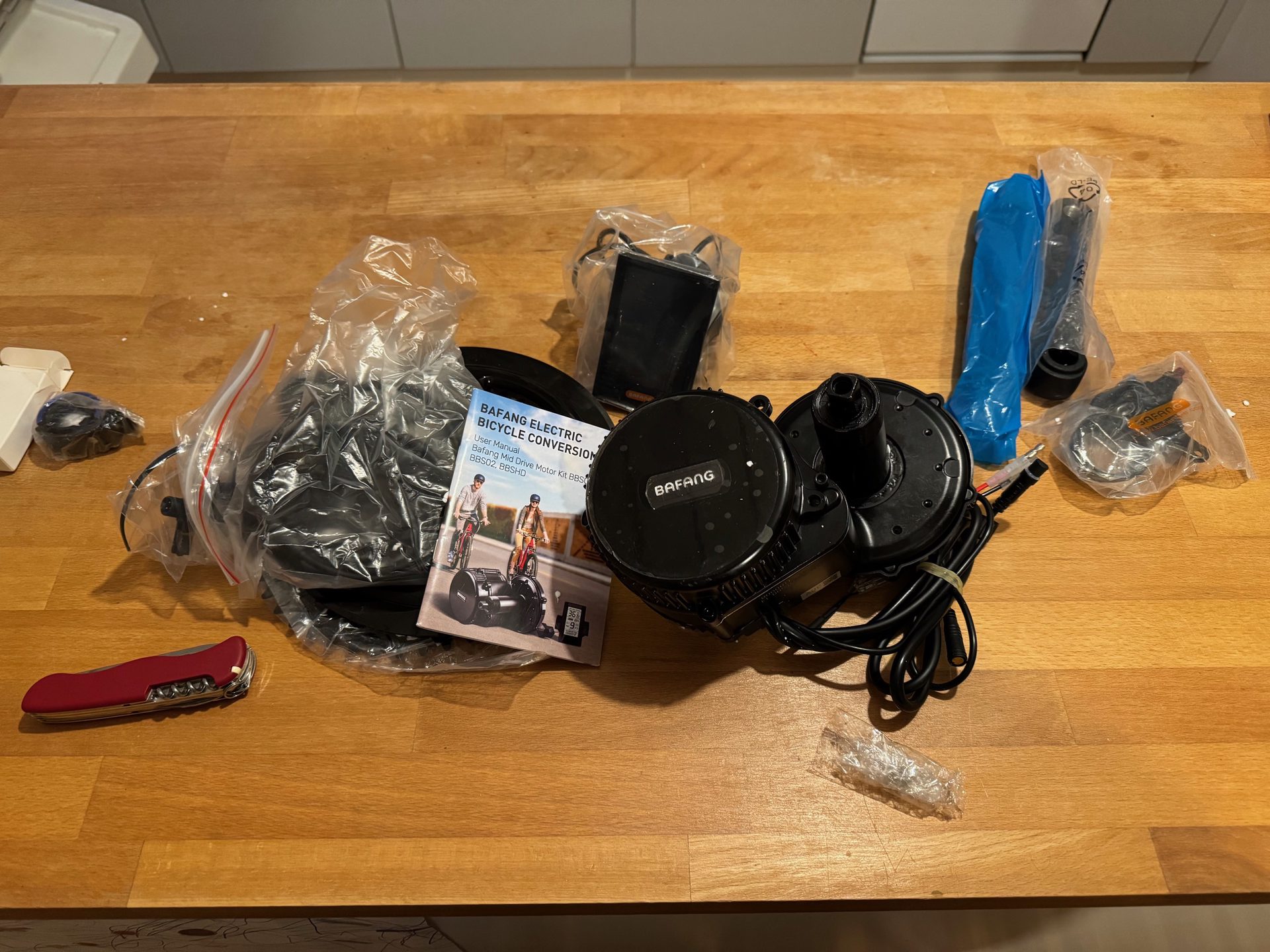 A Bafang BBS02 mid-drive motor, its manual, cables and bagged fittings laid out on a wooden kitchen table