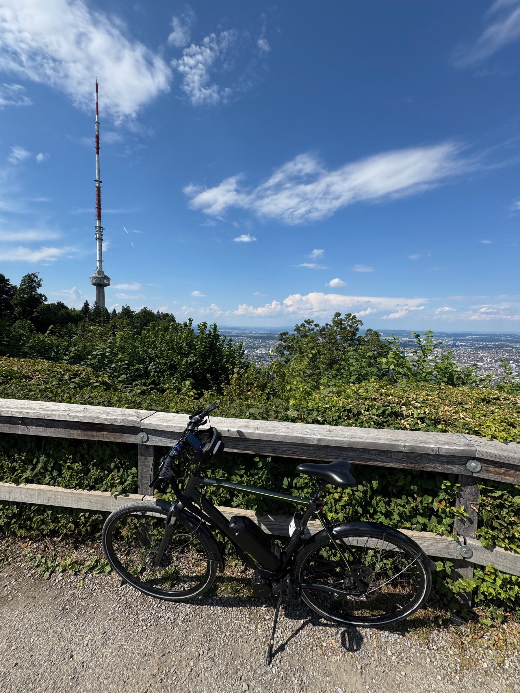 The bike leaning against a wooden fence at a hilltop viewpoint, a lattice TV tower on the skyline and a city hazy in the valley below