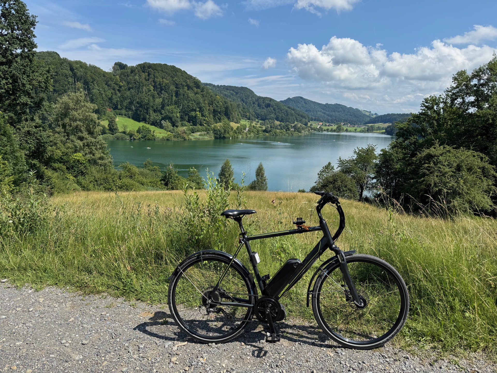 The converted bike parked on a gravel path above a lake, wooded hills behind it