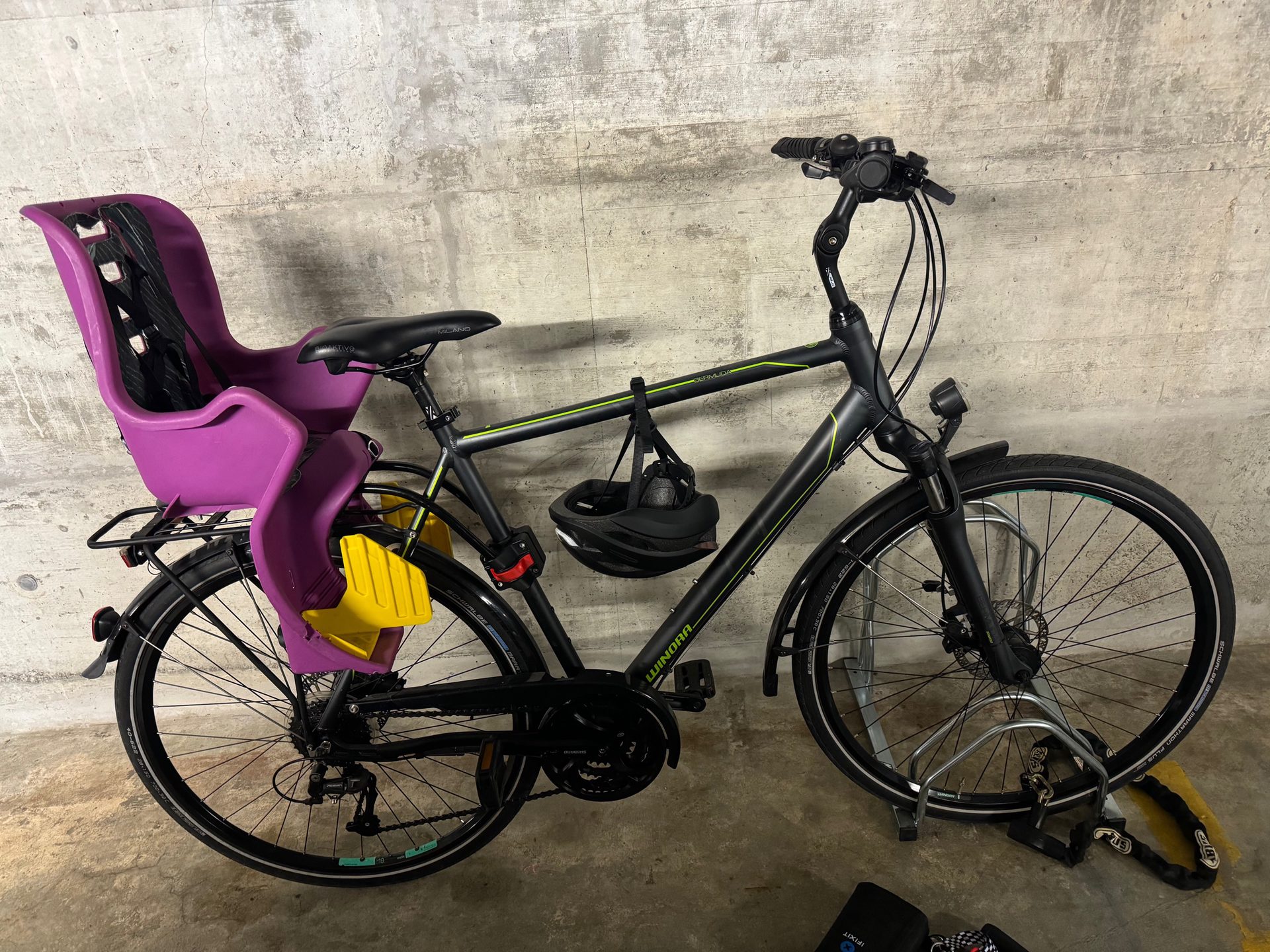 A stock black Winora Bermuda hybrid bike with mudguards, a rear rack and a purple child seat, parked against a concrete garage wall